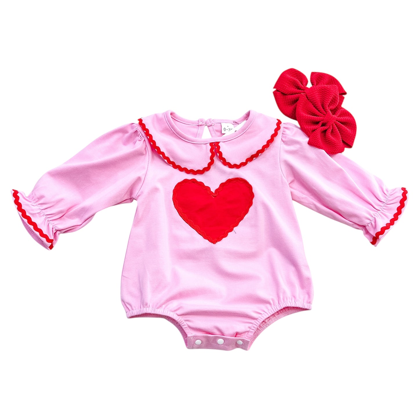 The Tiny Tot Bundles Classic Sweetheart Romper features long sleeves, red trim, a large red heart on the chest, and comes with a big red bow accessory near the neckline.