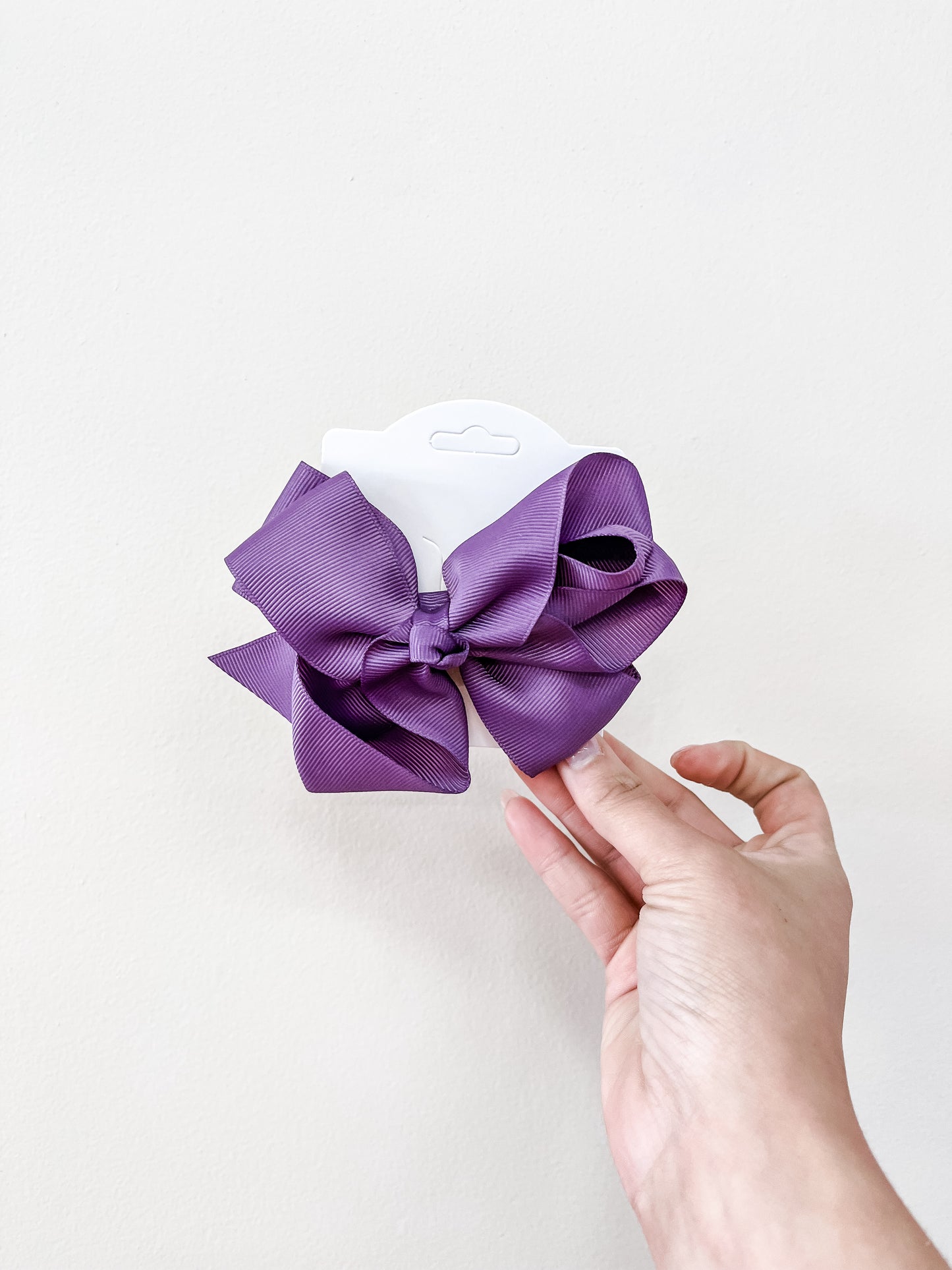 A hand holds the funteze Classic Boutique Bow in purple, attached to a white card, set against a plain light background.