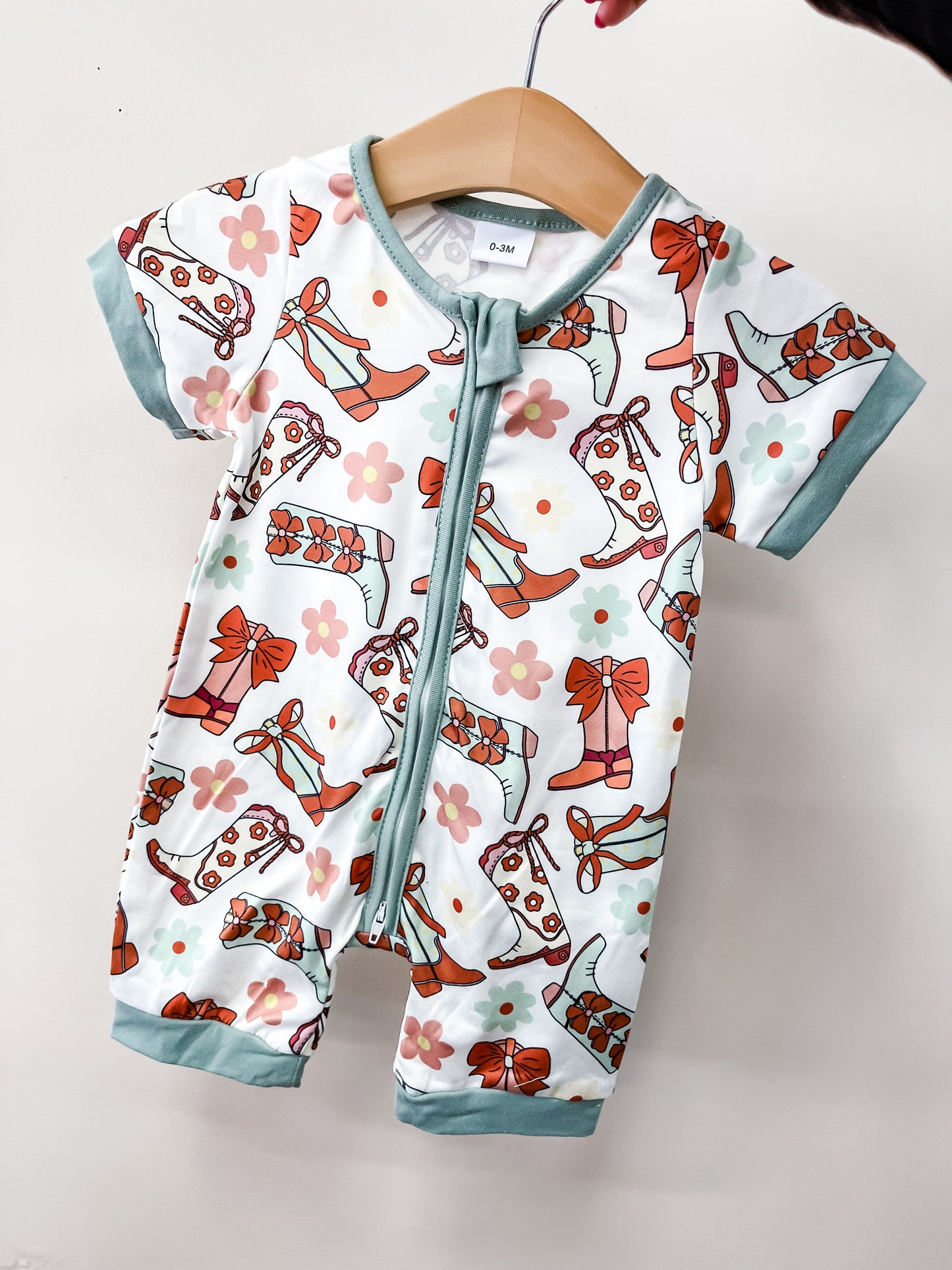 The Tiny Tot Bundles Spring Shower Romper features short sleeves, light blue trim, and a front zipper. Its colorful cowboy boots, flowers, and bows pattern stands out when displayed on a wooden hanger against a plain light background.
