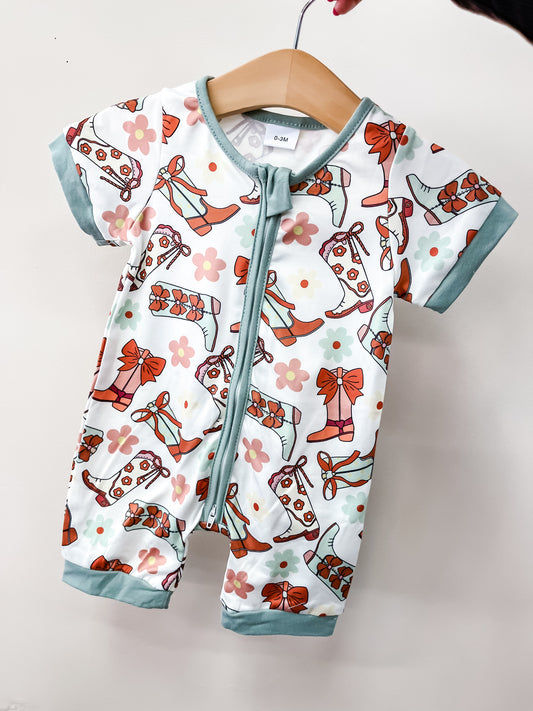 The Tiny Tot Bundles Spring Shower Romper features short sleeves, light blue trim, and a front zipper. Its colorful cowboy boots, flowers, and bows pattern stands out when displayed on a wooden hanger against a plain light background.