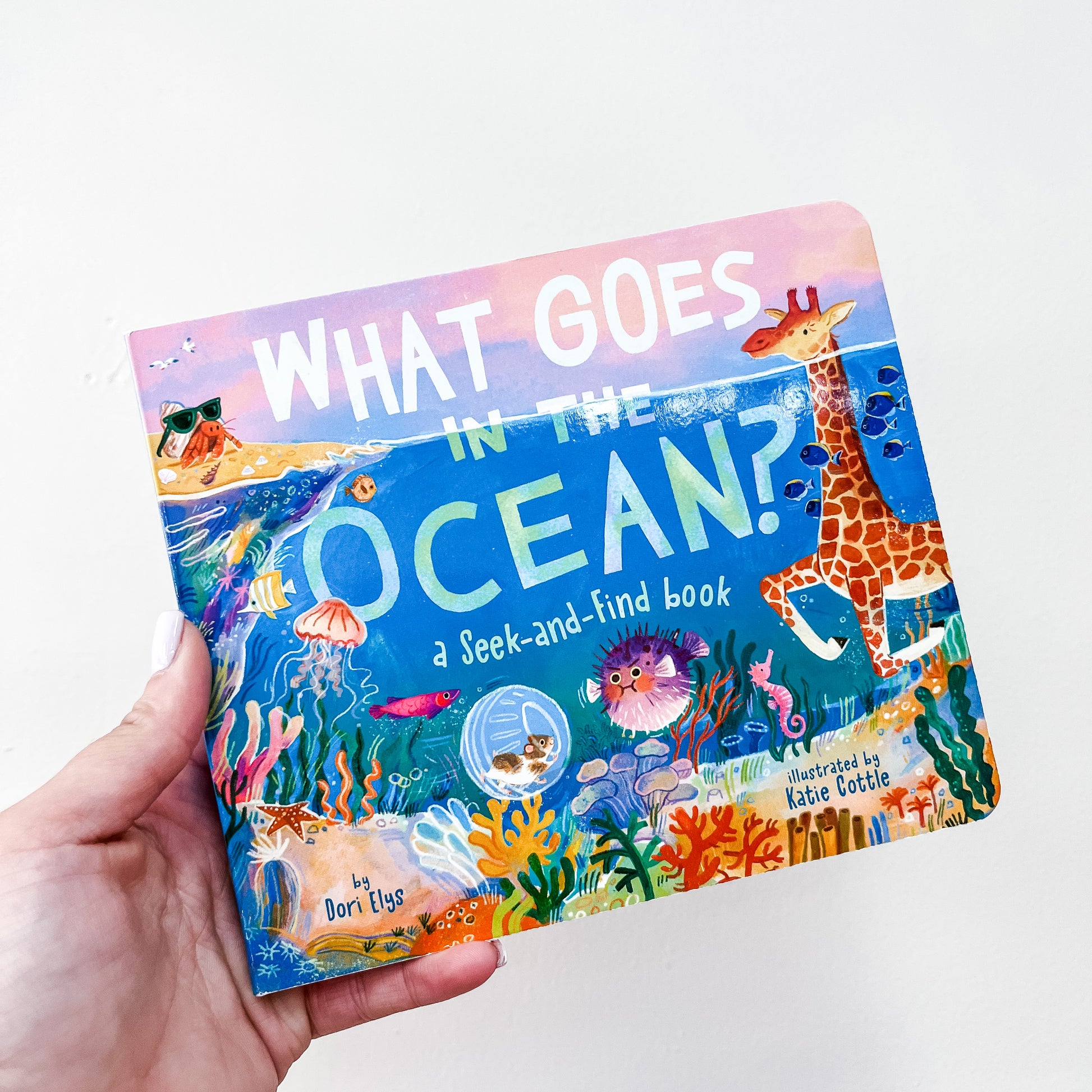 A hand holds "What Goes In The Ocean Book" by Simon & Schuster, a colorful children's seek-and-find with ocean animals and a giraffe on the cover.
