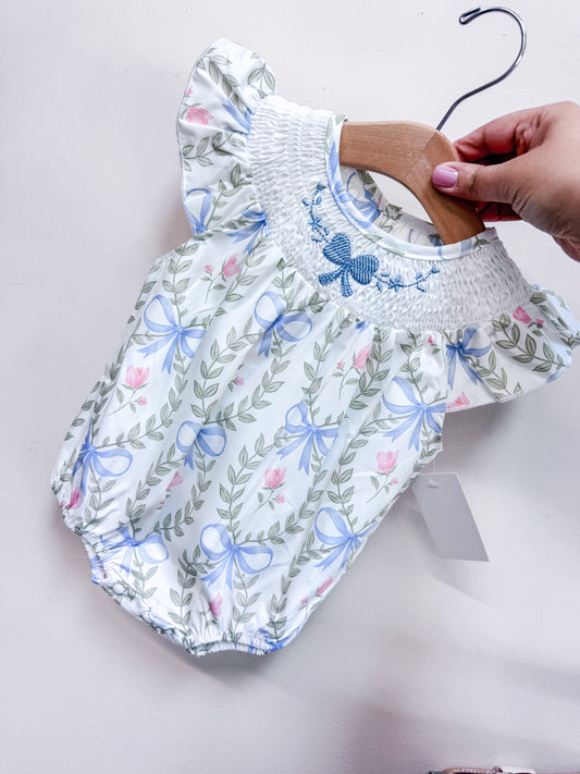 A hand holds the Sweet Garden Bow Romper by Tiny Tot Bundles on a wooden hanger. This white baby romper features a floral and blue bow pattern, short sleeves, and smocked neckline with embroidered blue flowers.