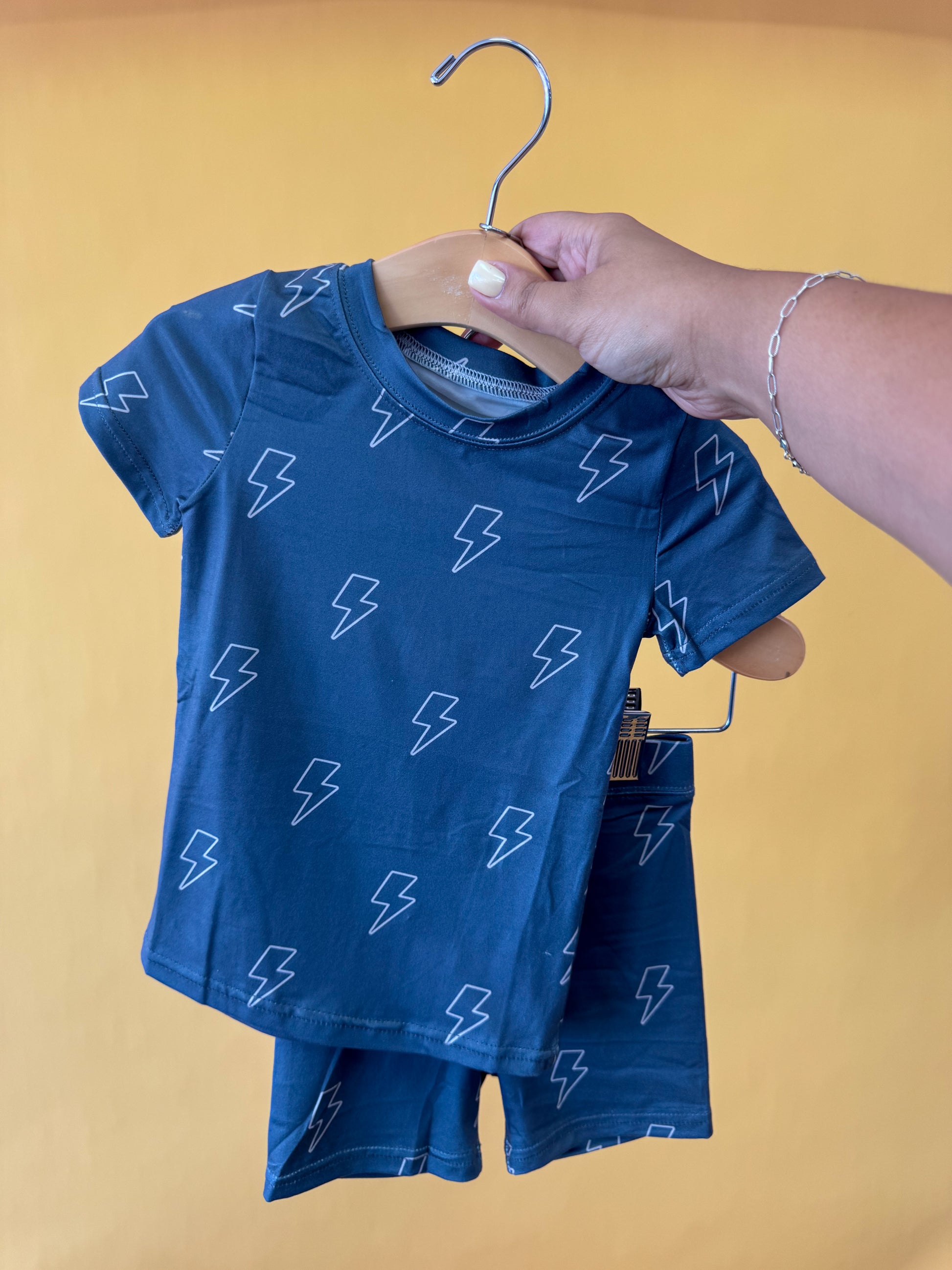 A hand holds up the Tiny Tot Bundles Storm Chaser Lounge Set—a blue children’s outfit with short sleeves and shorts, featuring white lightning bolt patterns—on a wooden hanger against a yellow background.