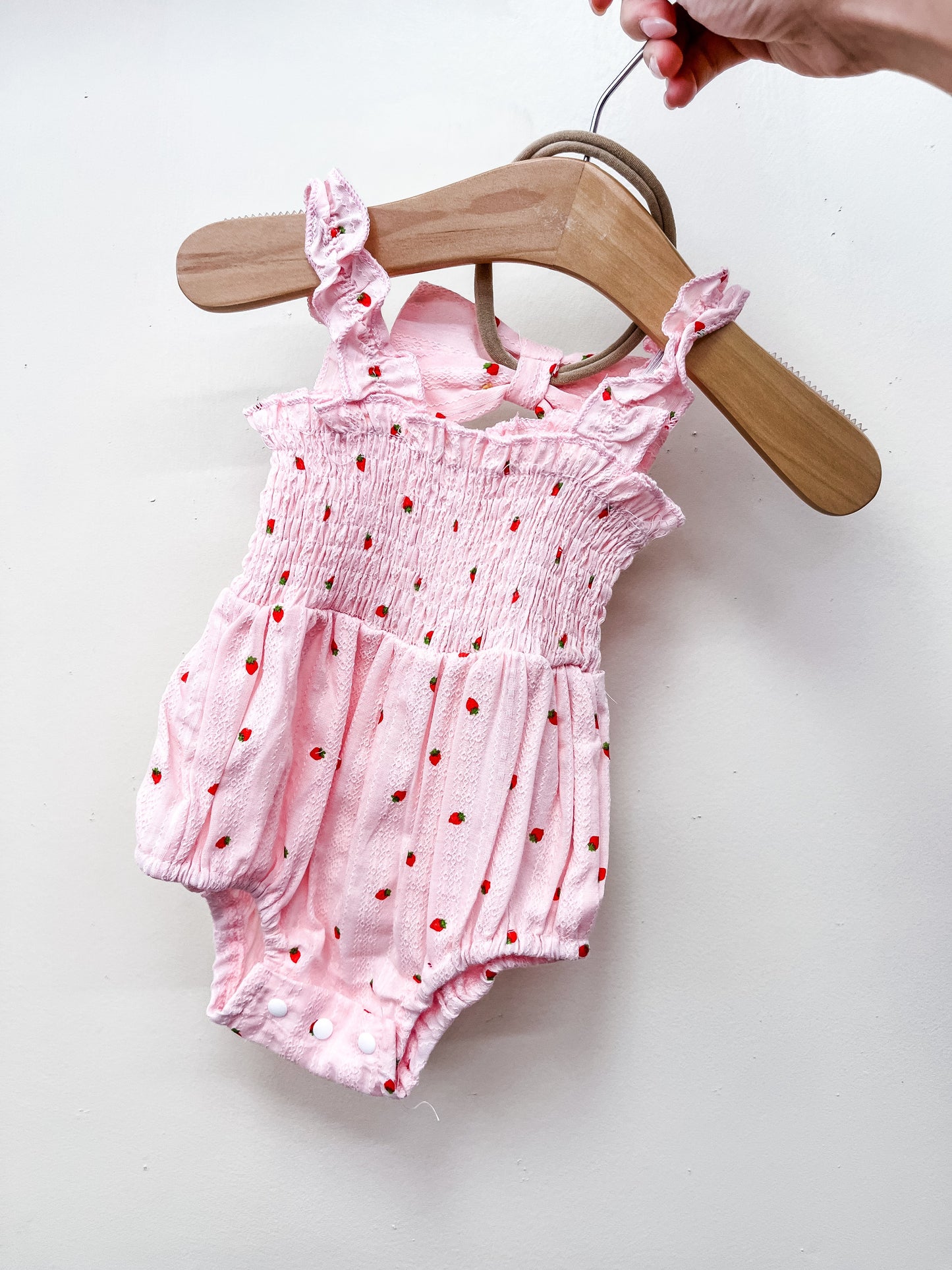 The Pink Strawberry Scrunch Romper by Tiny Tot Bundles, featuring ruffled straps, a smocked bodice, and red dot accents, is shown on a wooden hanger held by hand against a plain white background.