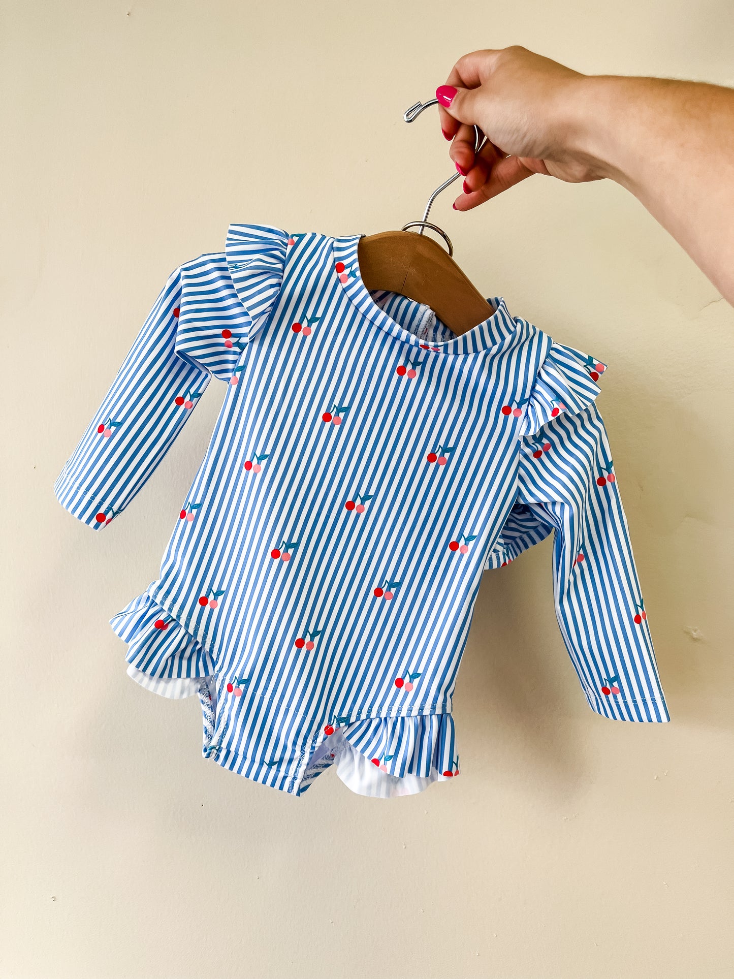 A hand holds a hanger displaying the Tiny Tot Bundles Cherry Stripe Swim Romper, featuring blue and white stripes, long sleeves, ruffle details, and small red cherries throughout.
