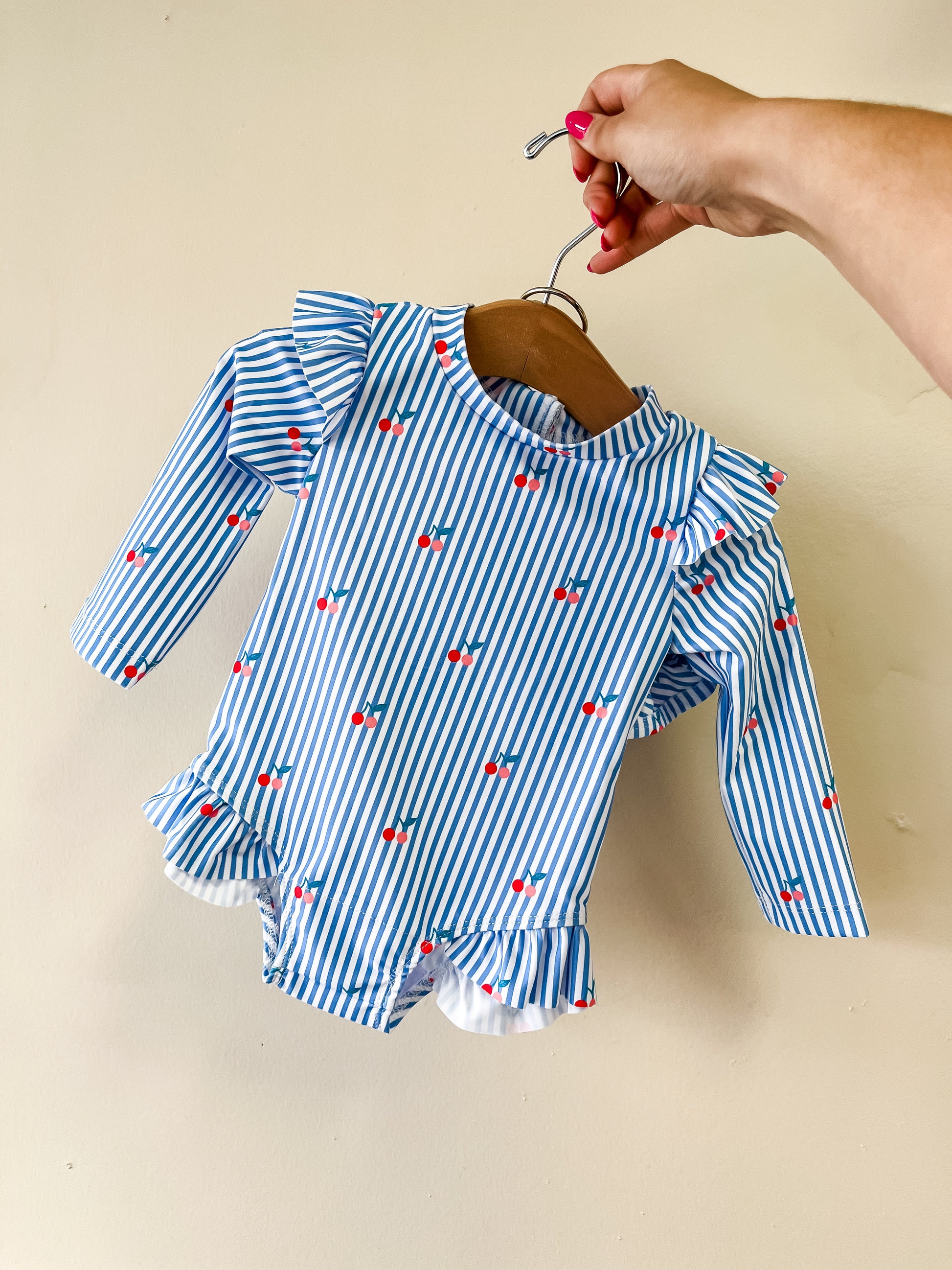 A hand holds a hanger displaying the Tiny Tot Bundles Cherry Stripe Swim Romper, featuring blue and white stripes, long sleeves, ruffle details, and small red cherries throughout.