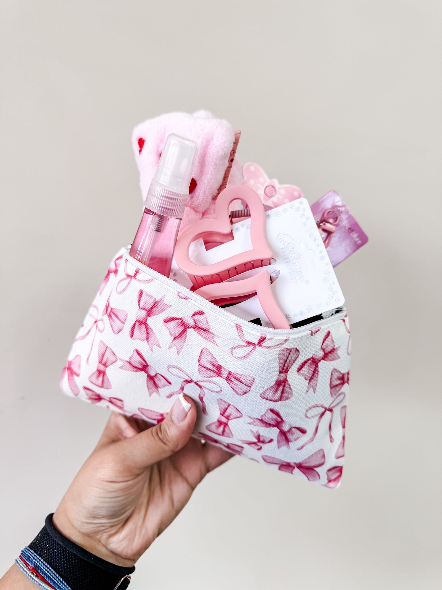 A hand holds the Tiny Tot Bundles Coquette Valentine’s Girl Bundle, a cosmetic pouch with pink bows, filled with pink and white items like a spray bottle, heart-shaped keychain, hair accessories, and a washcloth against a light background.