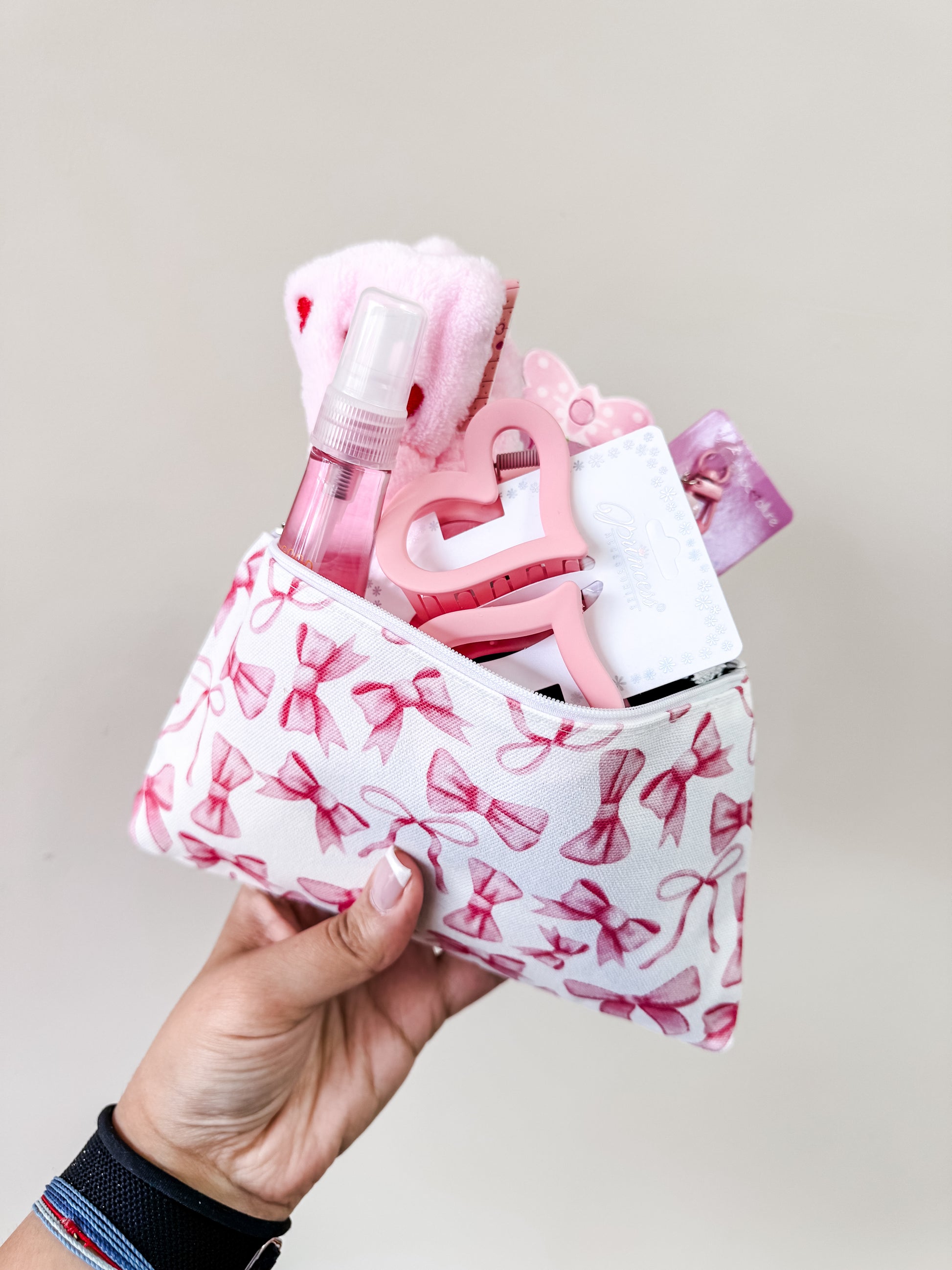 A hand holds the Tiny Tot Bundles Coquette Valentine’s Girl Bundle, a cosmetic pouch with pink bows, filled with pink and white items like a spray bottle, heart-shaped keychain, hair accessories, and a washcloth against a light background.