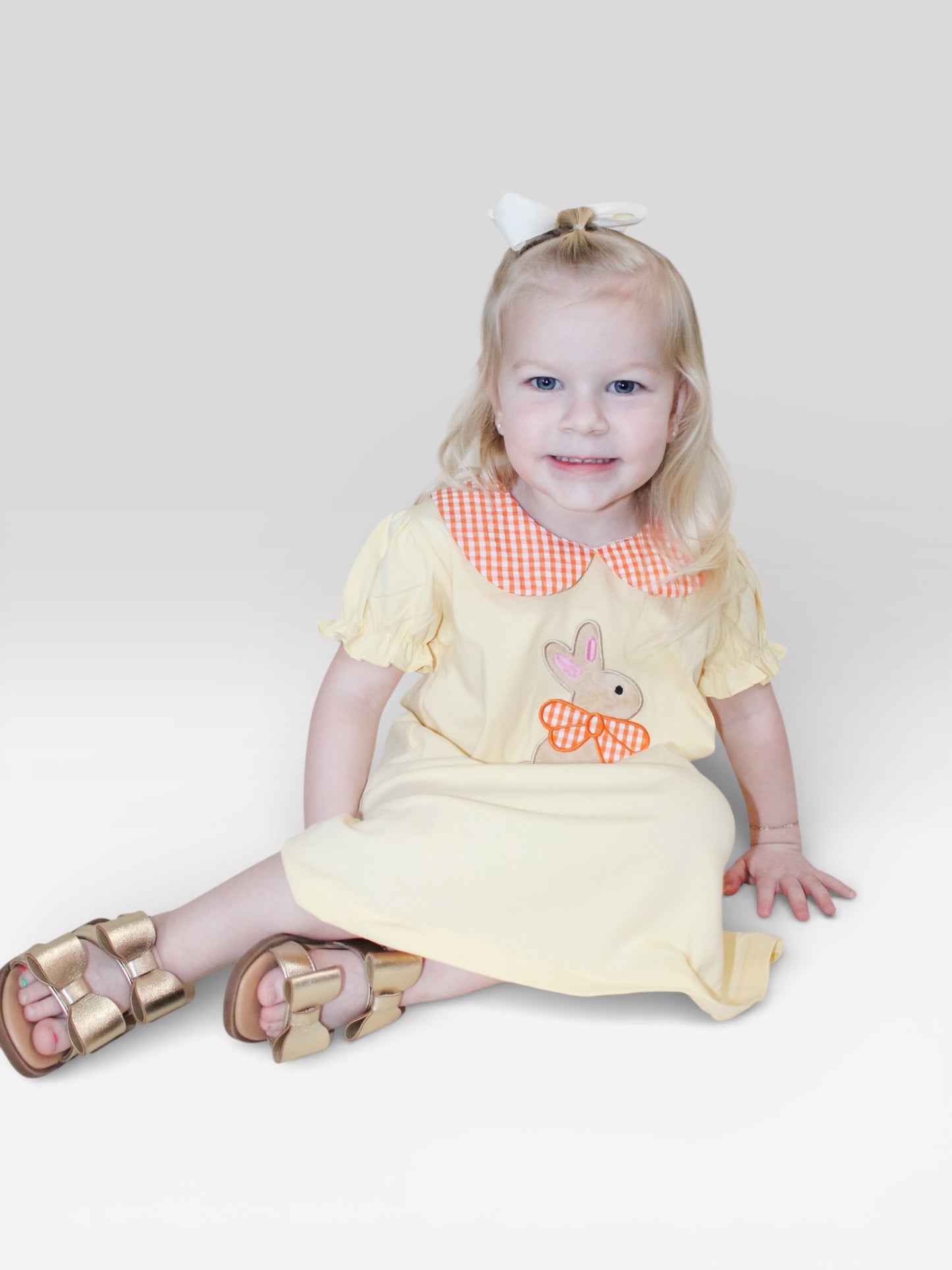A young blonde girl with a white bow smiles while sitting on the floor, wearing the Tiny Tot Bundles Buttercup Bunny Dress, featuring a gingham collar and bunny design. She pairs it with gold sandals against a plain light gray background.