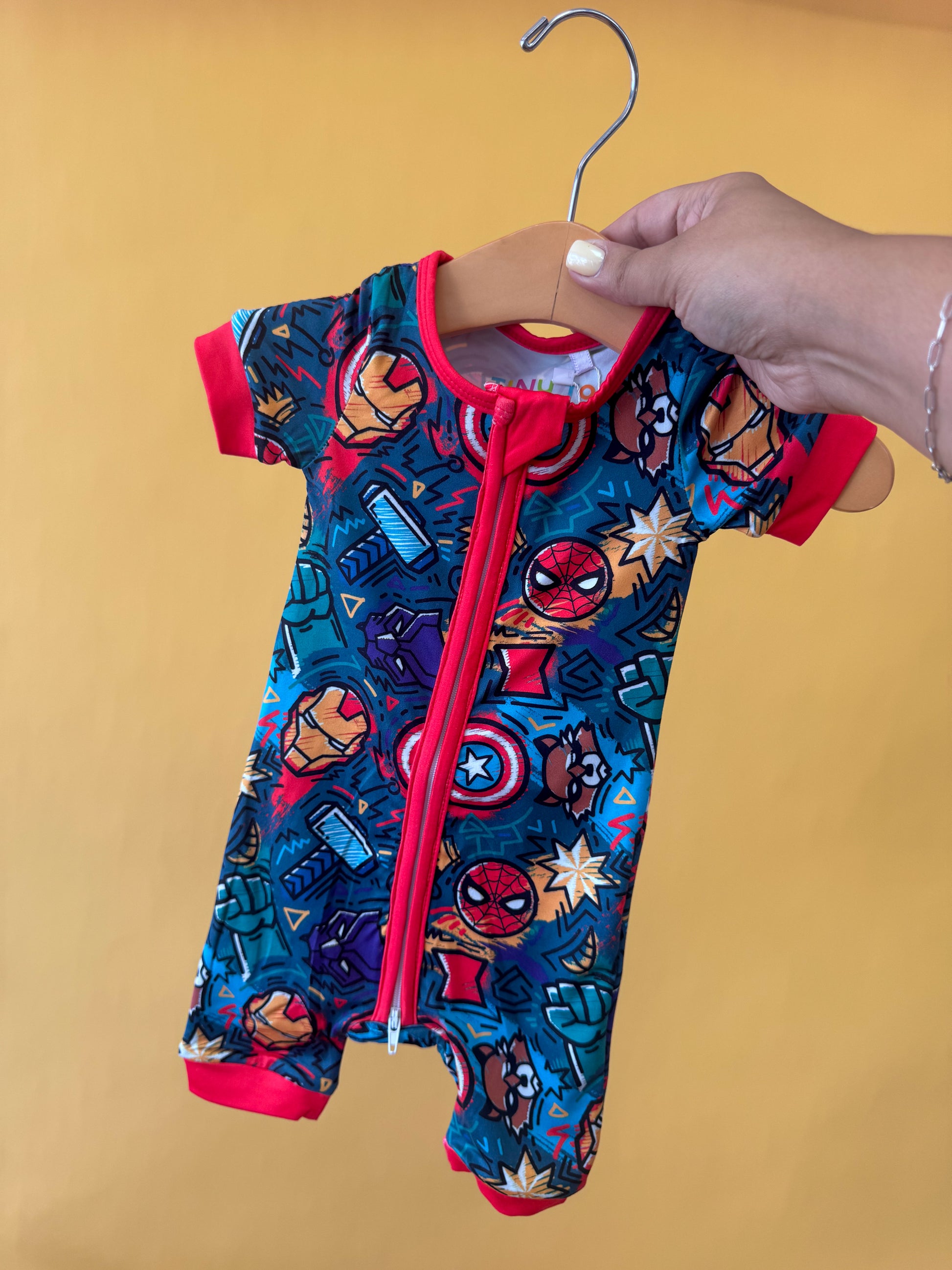 A hand holds the Tiny Tot Bundles Hero Headquarters Short Zippy, a colorful baby romper with red trim and a superhero pattern featuring masks and shields, displayed on a wooden hanger against a plain yellow background.