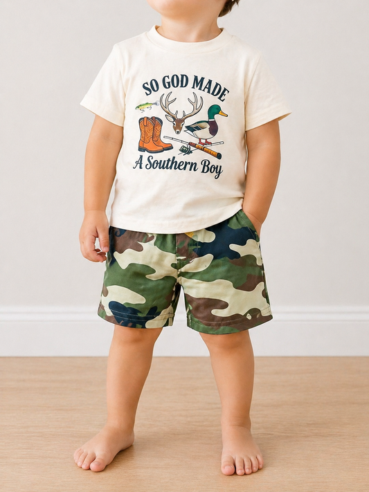 A young child stands barefoot indoors wearing the Southern Boy Camo Set from Tiny Tot Bundles—a cream tee with “So God Made A Southern Boy” and outdoor graphics, paired with green camo shorts.