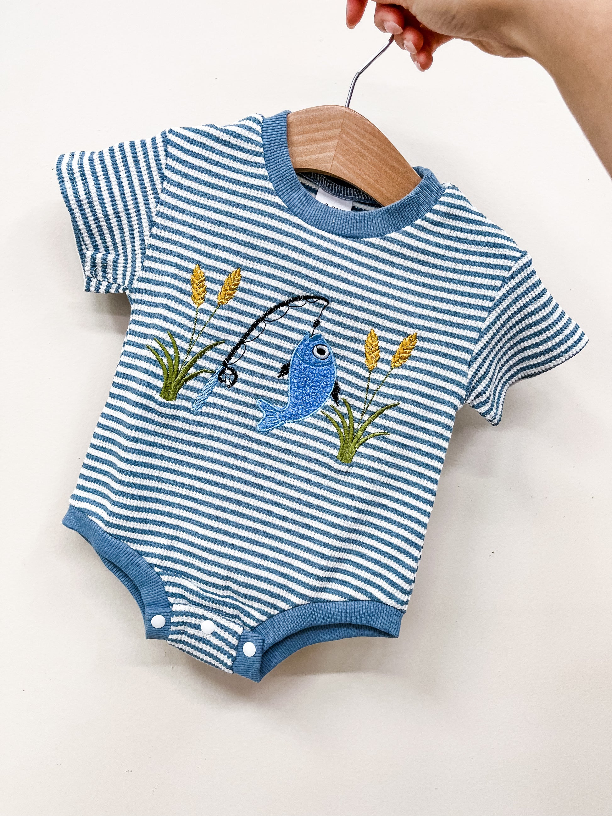 The Tiny Tot Bundles Fish Hook Stripe Bubble is a blue and white striped romper with blue trim and an embroidered fish-on-a-line among green cattails, shown on a wooden hanger held by a hand.