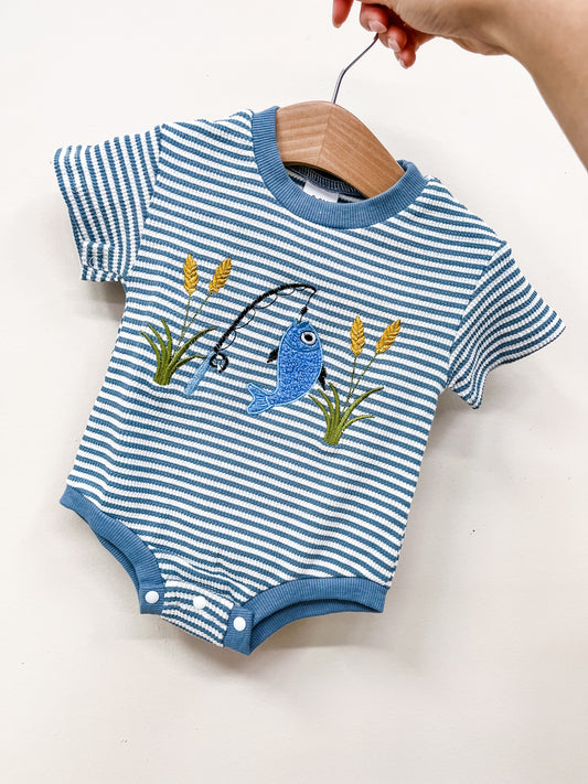 The Tiny Tot Bundles Fish Hook Stripe Bubble is a blue and white striped romper with blue trim and an embroidered fish-on-a-line among green cattails, shown on a wooden hanger held by a hand.