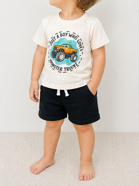 A young boy stands barefoot with hands in pockets, wearing the Tiny Tot Bundles Monster Truck Mania Set—cream t-shirt with a monster truck graphic and "Just a boy who loves monster trucks" text, paired with black shorts. Face not shown.