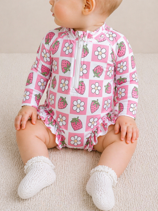 A baby in a Tiny Tot Bundles Strawberry Check Swimsuit sits sideways on a light carpet, also wearing white knitted socks.