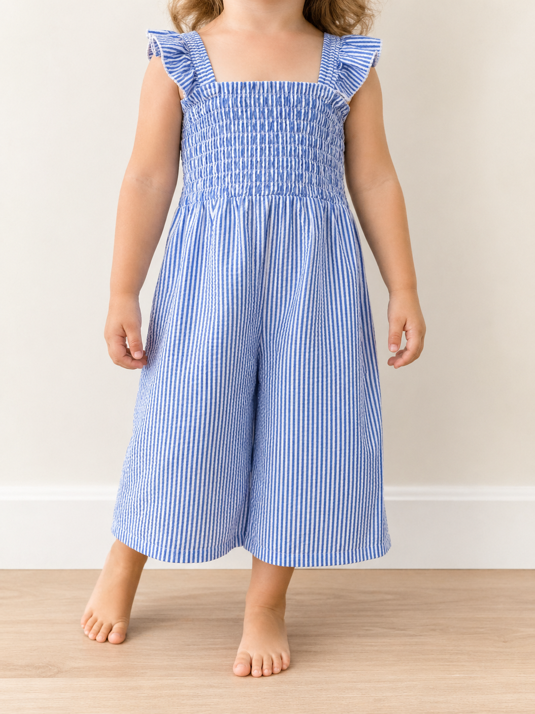 A young child wears the Tiny Tot Bundles Blue Stripe Smocked Jumpsuit, featuring sleeveless, blue and white stripes with ruffled shoulder straps, while standing barefoot on a wooden floor against a light-colored wall.