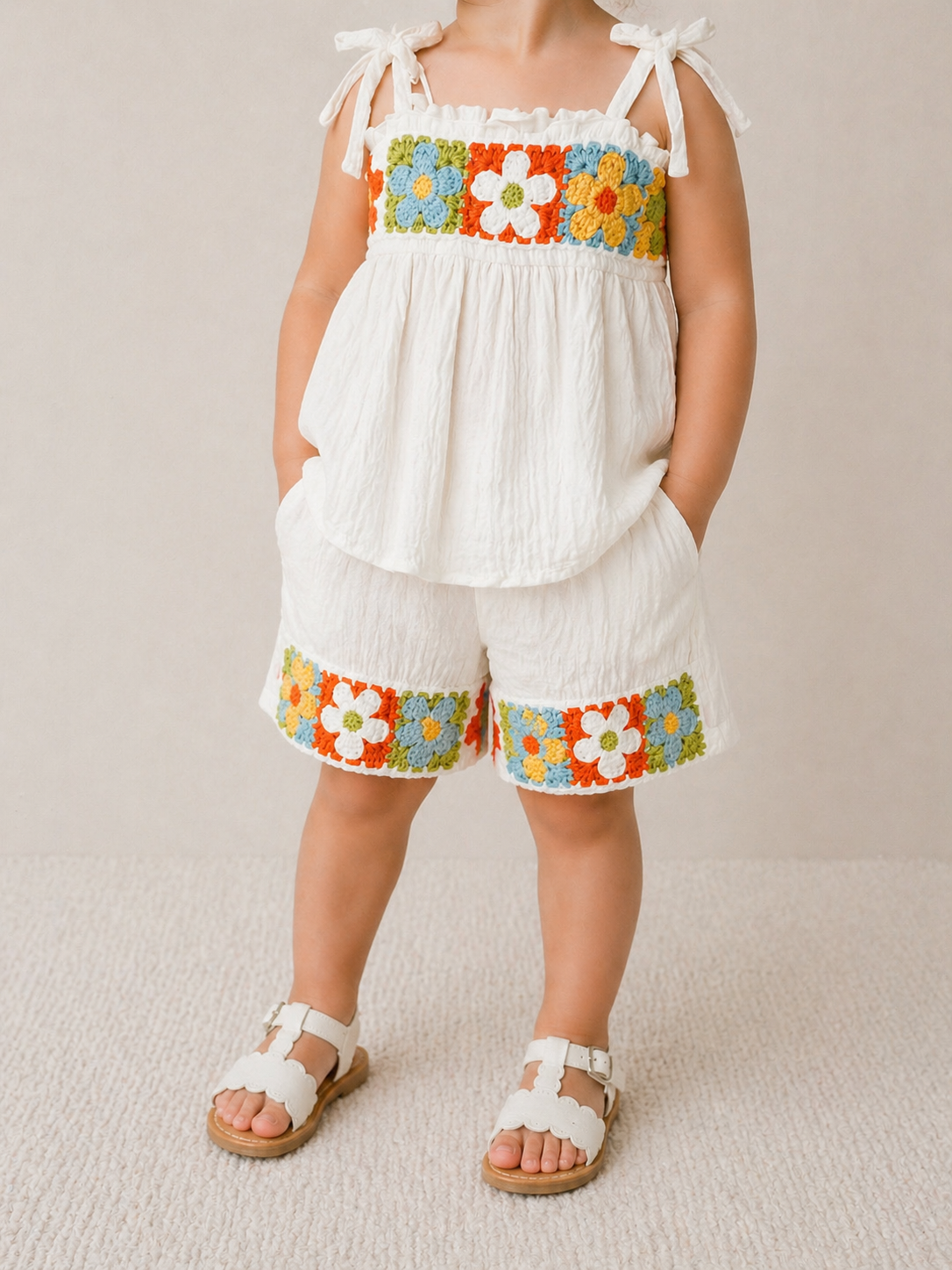 A young child wears the Sunshine Crochet Set by Tiny Tot Bundles—a white sleeveless top and shorts featuring colorful floral embroidery—paired with white sandals. The child's face is not shown.