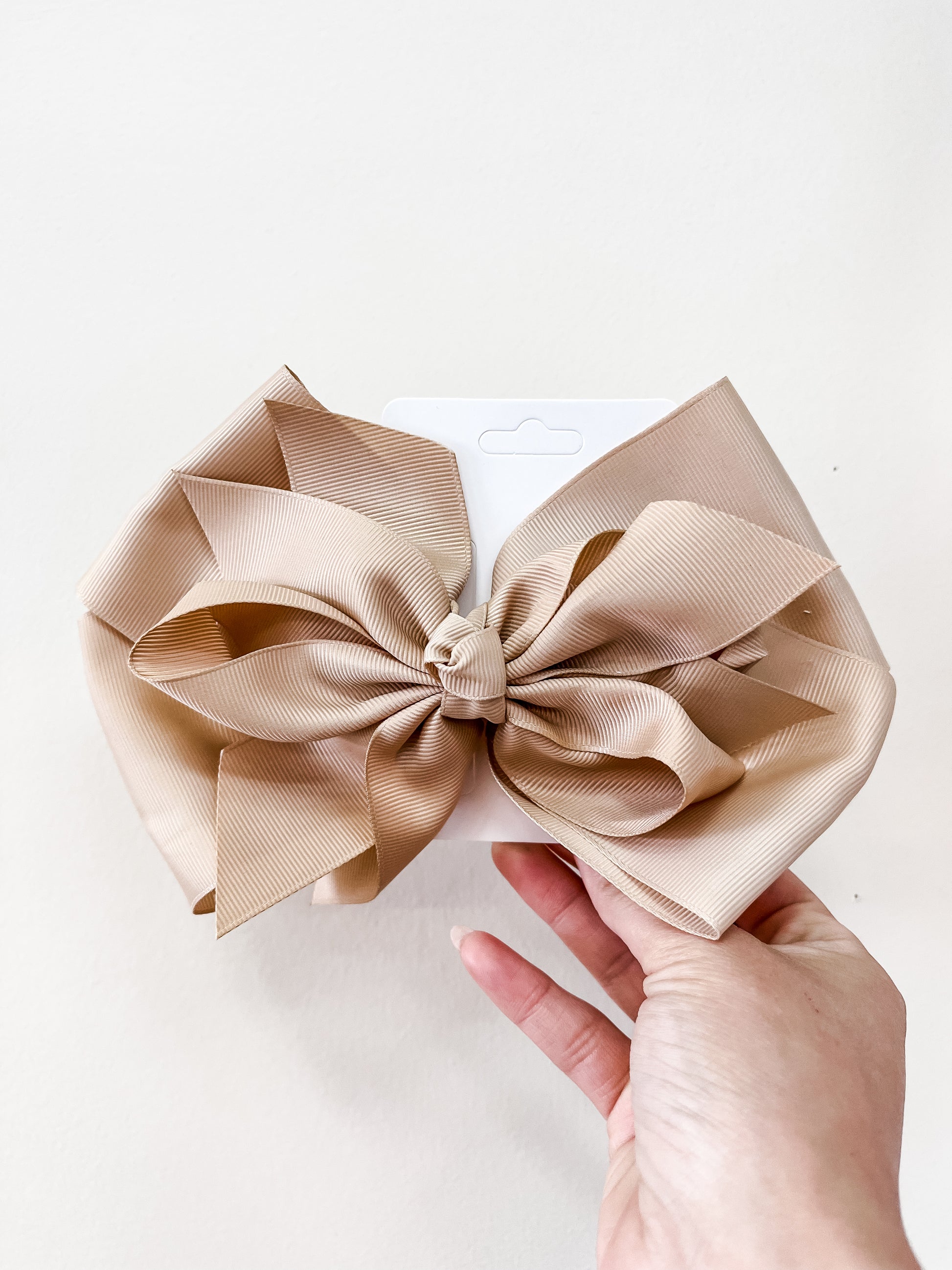 A hand holds an Oversized Pastel Bow by funteze, in beige, attached to a white card against a plain light background.