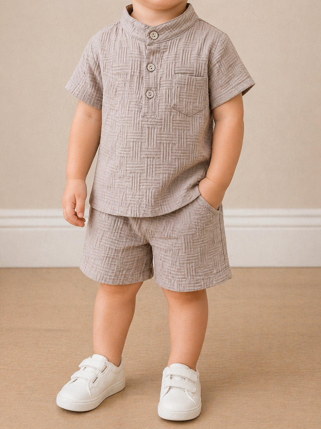 A toddler models the Tiny Tot Bundles Textured Boy Button Up Set—featuring a beige short-sleeve shirt with three buttons and matching shorts—paired with white sneakers, standing against a neutral background.