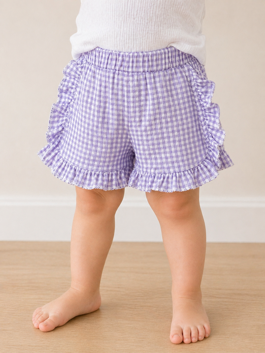 A young child stands barefoot on a wooden floor, wearing Tiny Tot Bundles' Picnic Day Gingham Shorts in purple with ruffled edges and a white top. The image is cropped to show only the child from the waist down.