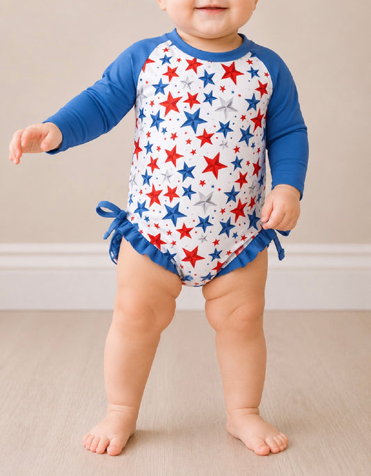 A baby stands on a light wood floor wearing the Tiny Tot Bundles Stars Swimsuit—a long-sleeve romper with blue sleeves, a white body with red and blue stars, blue ruffles, and side bows at the hips.