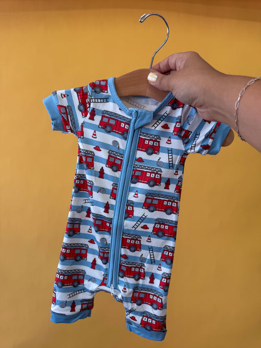 A hand holds the Tiny Tot Bundles Rescue Ready Short Zippy, a baby romper with blue trim featuring red fire trucks, cones, and ladders, against a plain orange background.