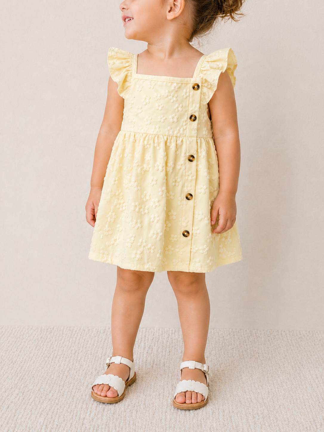 A young child stands on a light carpet, looking to the side, wearing Tiny Tot Bundles’ Lemon Blossom Dress—a light yellow, textured dress with ruffled sleeves and side buttons—paired with white sandals.