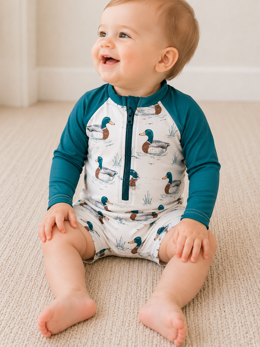 A smiling baby sits on a carpet in the Tiny Tot Bundles Little Duckling Swim Romper, featuring teal sleeves and a duck pattern, looking slightly to the side with hands resting on their legs.