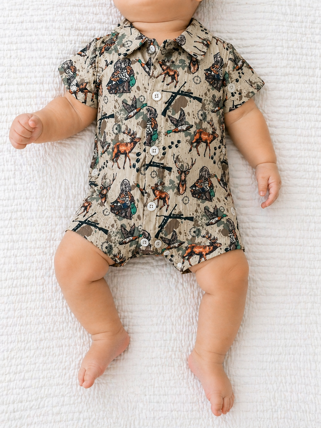 A baby lies on a white textured blanket wearing the Tiny Tot Bundles Little Hunter Bubble, a short-sleeve, button-up romper with a hunting-themed print in earthy tones. The baby's face is not visible.