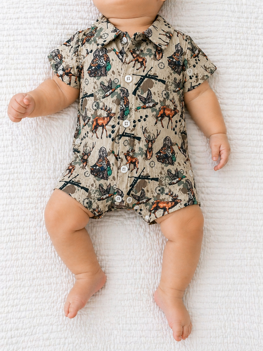 A baby lies on a white textured blanket wearing the Tiny Tot Bundles Little Hunter Bubble, a short-sleeve, button-up romper with a hunting-themed print in earthy tones. The baby's face is not visible.