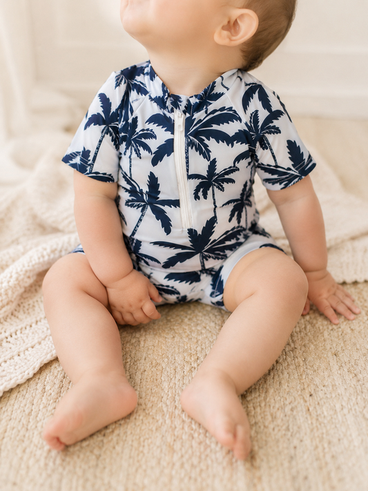 A baby in the Tiny Tot Bundles Palm Coast Swimsuit sits on a light woven rug, looking up with hands on their legs. A knitted blanket is partially visible beside them.