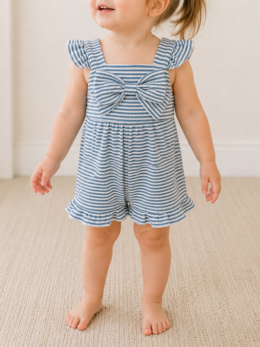 A young child indoors wears the Bluebell Breeze Bow Romper by Tiny Tot Bundles, featuring blue and white stripes, ruffled sleeves, and a large chest bow. The child’s hair is in a ponytail; the room has neutral carpet and light walls.