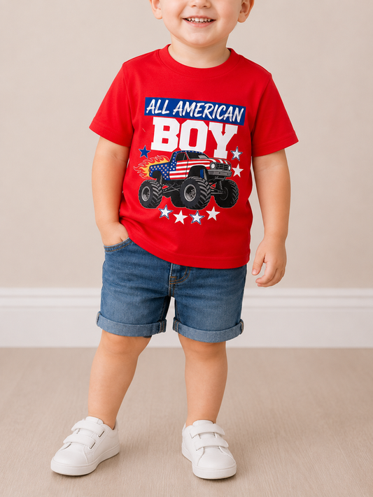 A smiling young child wears the Tiny Tot Bundles All-American Boy Tee featuring an “All American Boy” text and stars-and-stripes monster truck graphic, paired with denim shorts and white sneakers, standing indoors against a light background.