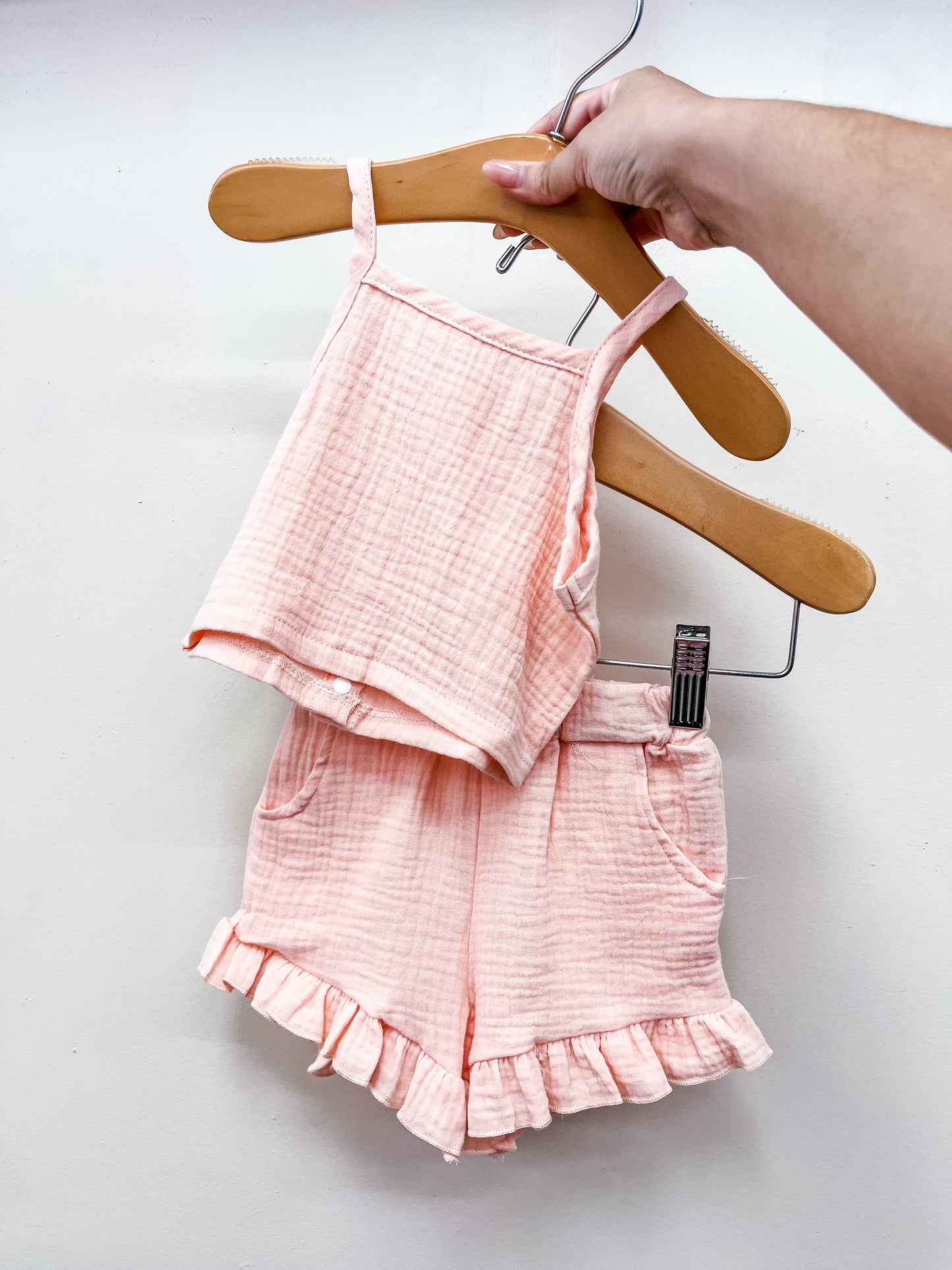 A hand holds up the Tiny Tot Bundles Pale Pink Linen Set—sleeveless top and matching shorts with ruffled hems—in soft, textured fabric, shown against a plain white background.