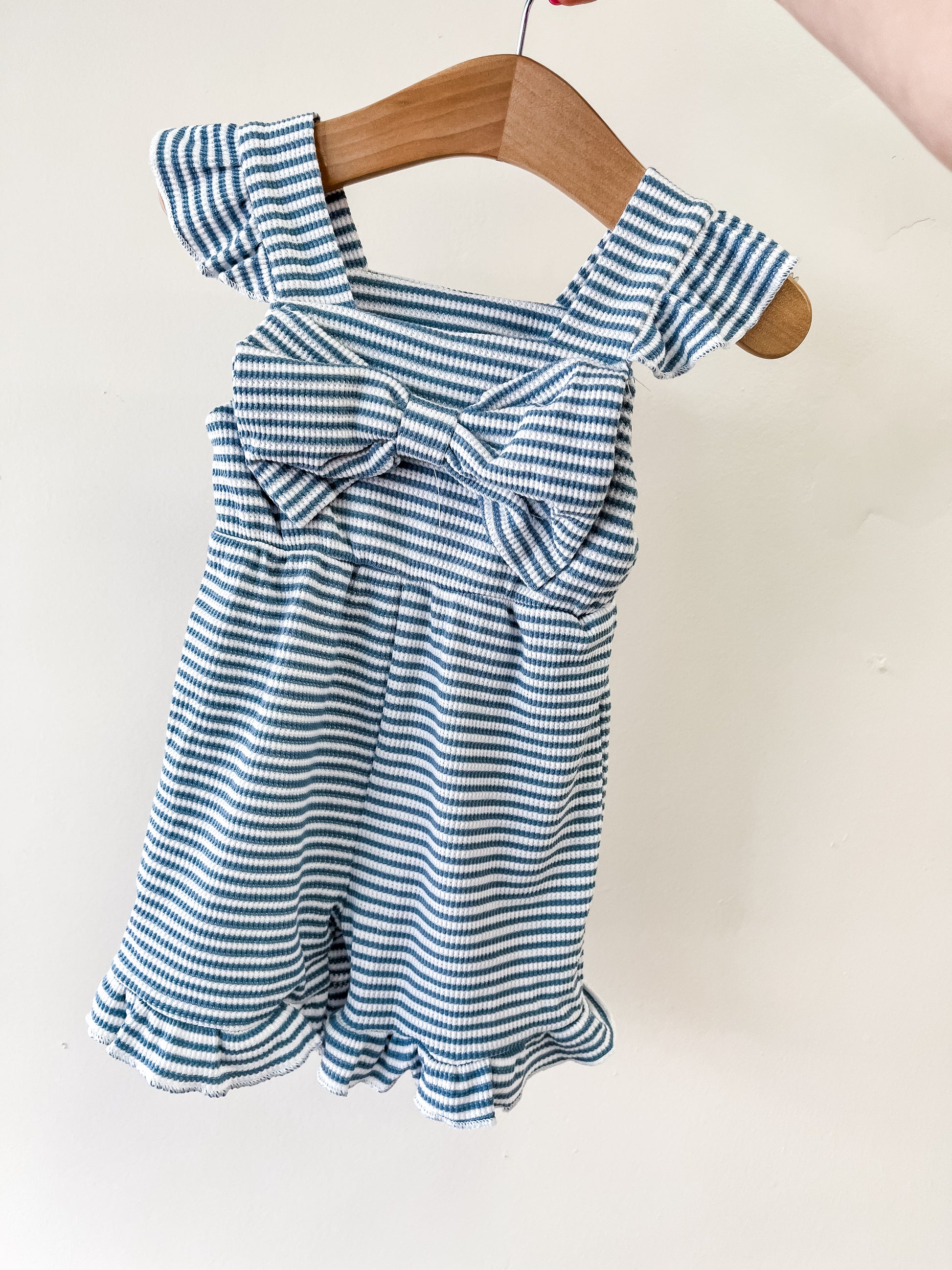 The Bluebell Breeze Bow Romper by Tiny Tot Bundles features blue and white stripes, ruffled sleeves and legs, a gathered waist, and a big chest bow, displayed on a wooden hanger against a plain white background.