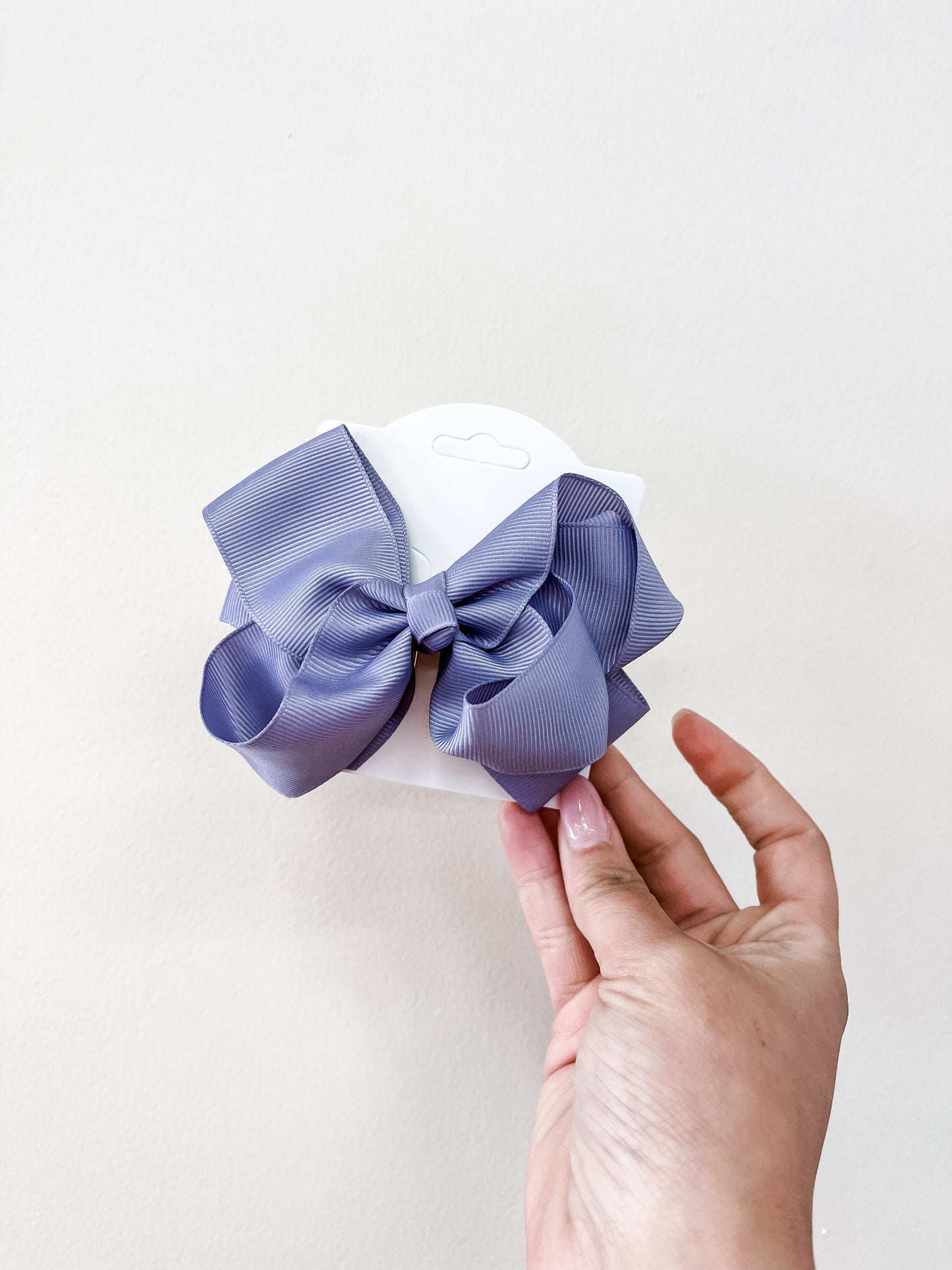 A hand with light pink nail polish holds a white card featuring the funteze Classic Boutique Bow—large and neatly tied in lavender—set against a plain off-white background.