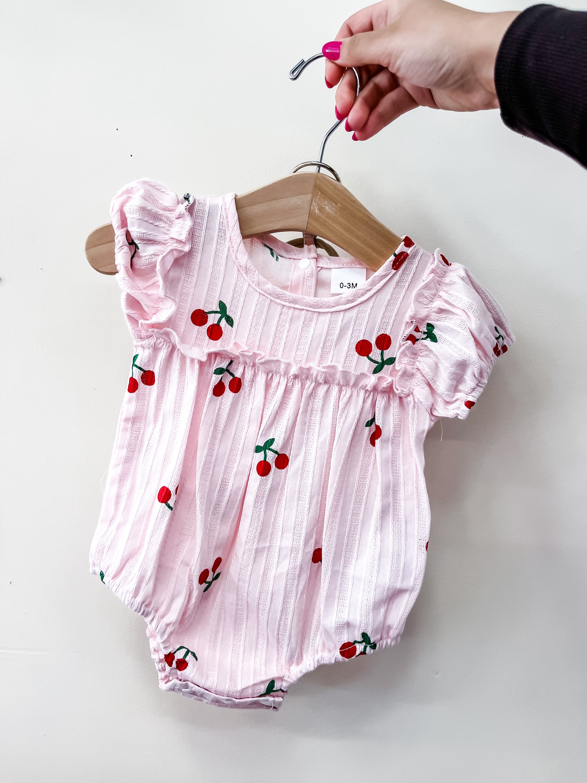 A hand holds a hanger with the Tiny Tot Bundles Cherry Blossom Bubble—a pink baby romper with a cherry pattern, ruffled sleeves, and ribbed texture—displayed against a white background.
