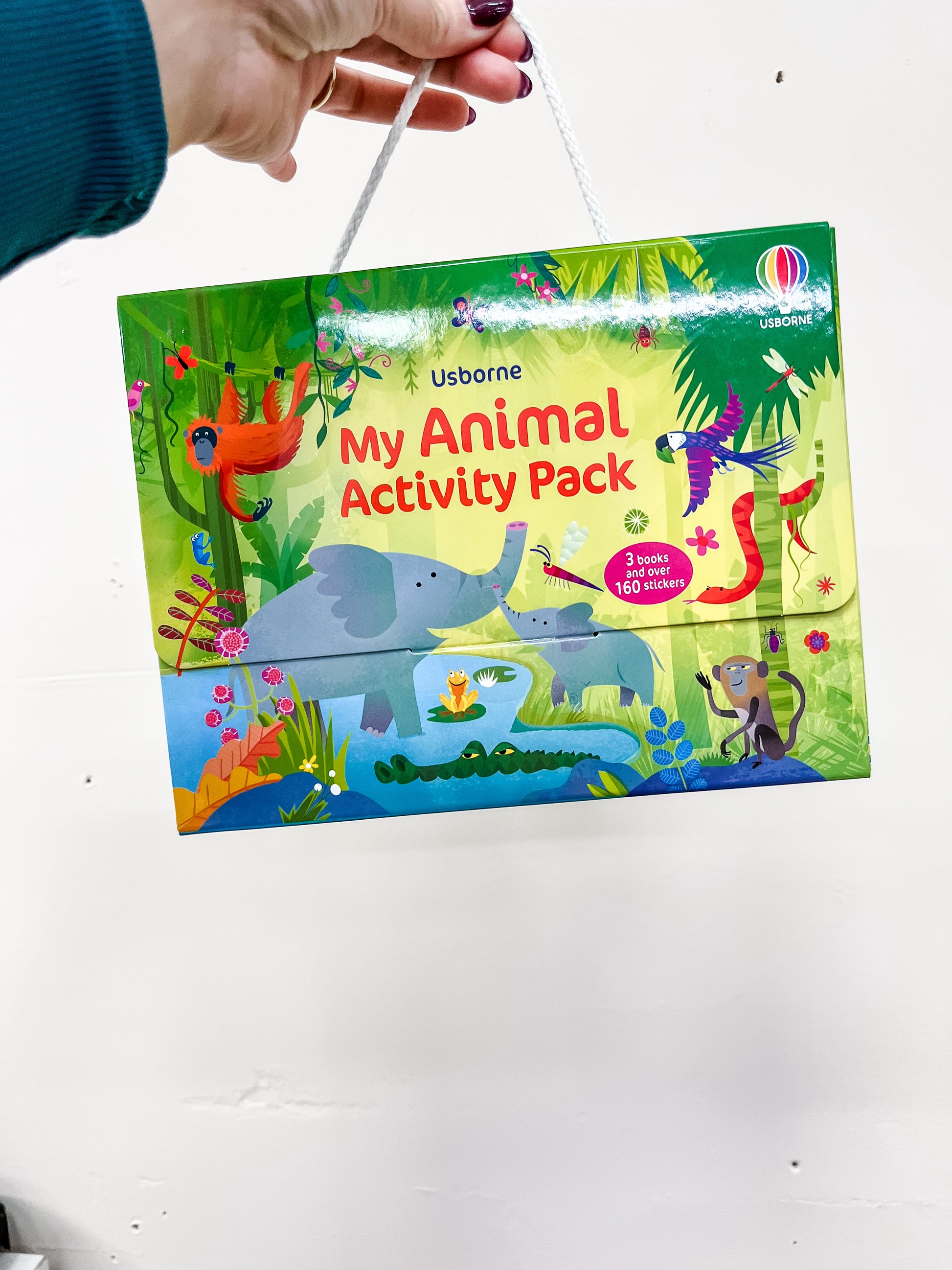A hand holds up a Harper Collins Animals Activity Pack Book with colorful jungle illustrations of animals like an elephant, monkey, parrot, crocodile, and snake.