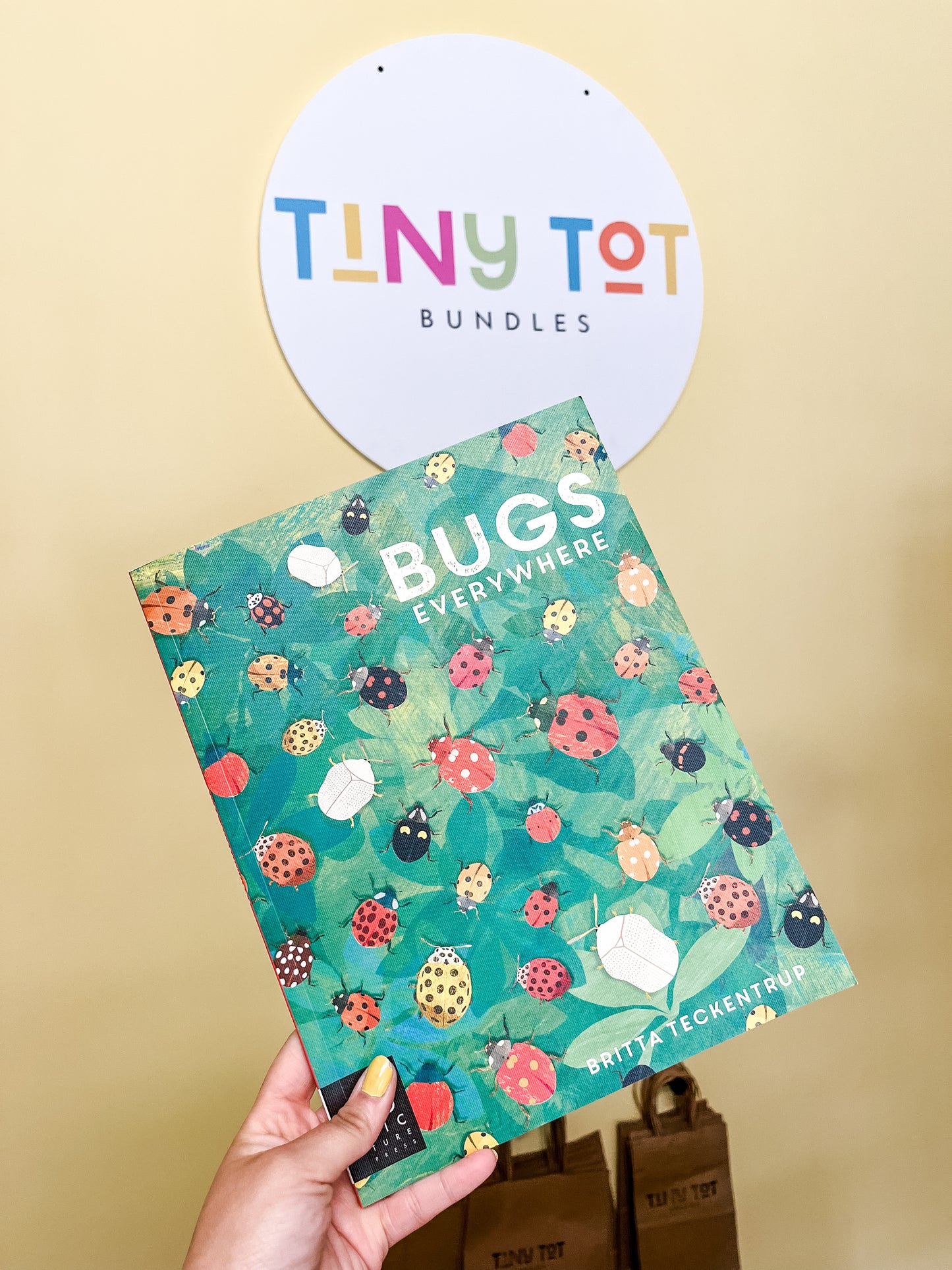 A hand holds the Bugs Everywhere Books by Tiny Tot Bundles, featuring a cover with colorful bug illustrations. In the background, a circular sign displays the Tiny Tot Bundles brand on a yellow wall.