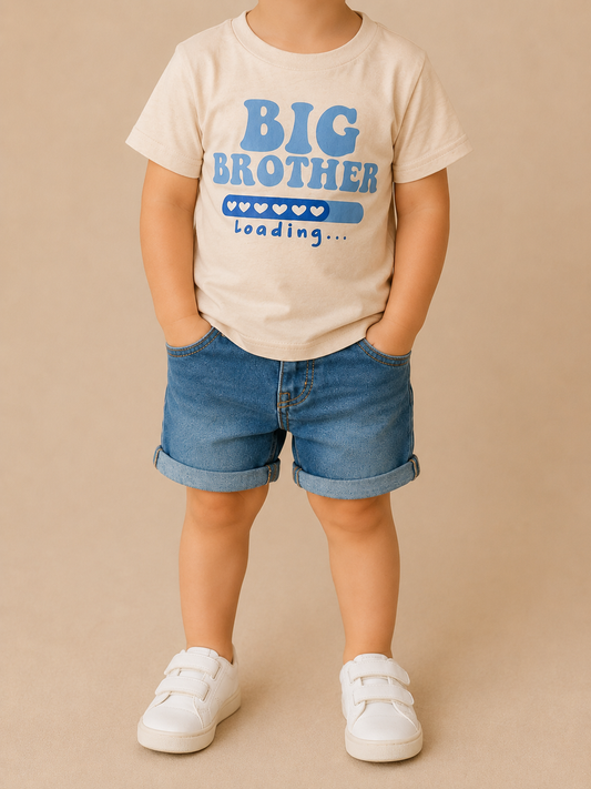 A young child wears the Big Brother Loading Tee by Tiny Tot Bundles, styled with denim shorts and white sneakers, standing against a beige background.