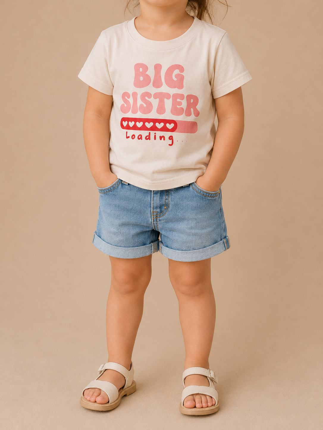 A young child stands with hands in pockets, modeling the Tiny Tot Bundles Big Sister Loading Tee with heart design, denim shorts, and white sandals against a beige background. The child's face is not shown.