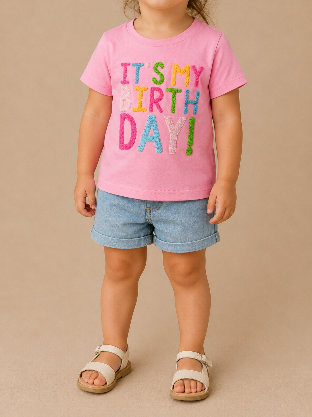A young child wears the Tiny Tot Bundles It's My Birthday Tee, featuring colorful lettering, paired with denim shorts and beige sandals, standing against a plain beige background.