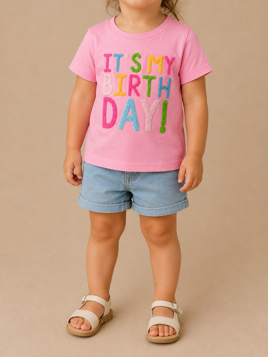 A young child wears the Tiny Tot Bundles It's My Birthday Tee, featuring colorful lettering, paired with denim shorts and beige sandals, standing against a plain beige background.