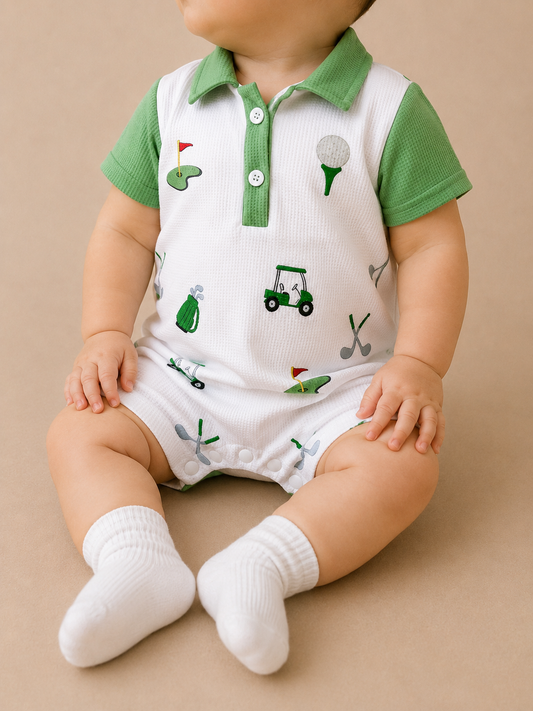 A baby in the Tiny Tot Bundles Golf Cart Adventures Romper, featuring green and white golf-themed prints, sits on a neutral background wearing white socks and looking to the side with hands on their legs.