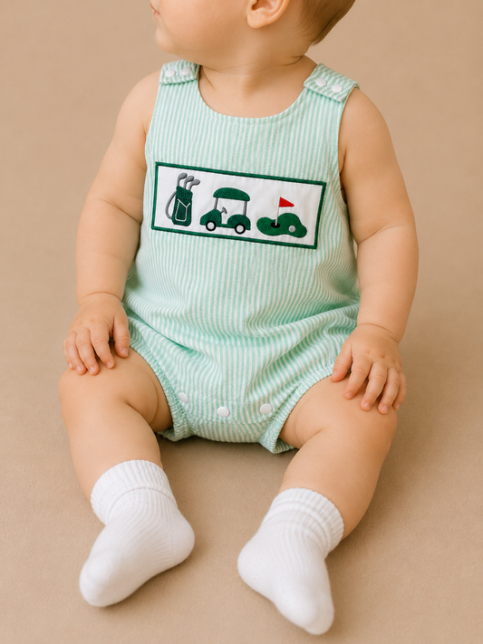 A baby sits on a beige surface wearing Tiny Tot Bundles’ Little Golfer Bubble, a green and white striped romper with embroidered golf bag, golf cart, and flag designs, paired with white socks.