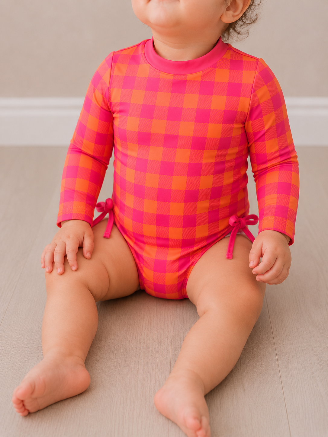 A toddler sits on a light wooden floor in Tiny Tot Bundles’ Sunset Splash Check Swimsuit, a pink and orange checkered long-sleeve style with small bows at the hips. The child’s face is partially out of frame.