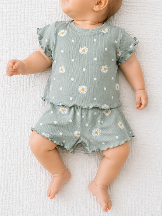 A baby lies on a white textured blanket, wearing the Tiny Tot Bundles Minty Meadow Set: a green short-sleeved top and matching shorts with white polka dots and daisy flower prints.