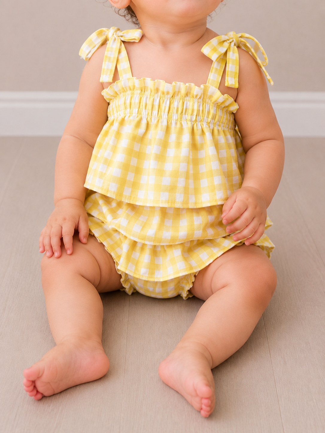 A baby in the Tiny Tot Bundles Sunny Picnic Gingham Set—yellow and white, sleeveless with ruffles and shoulder ties—sits on a light wooden floor against a neutral wall, their face partially out of frame.