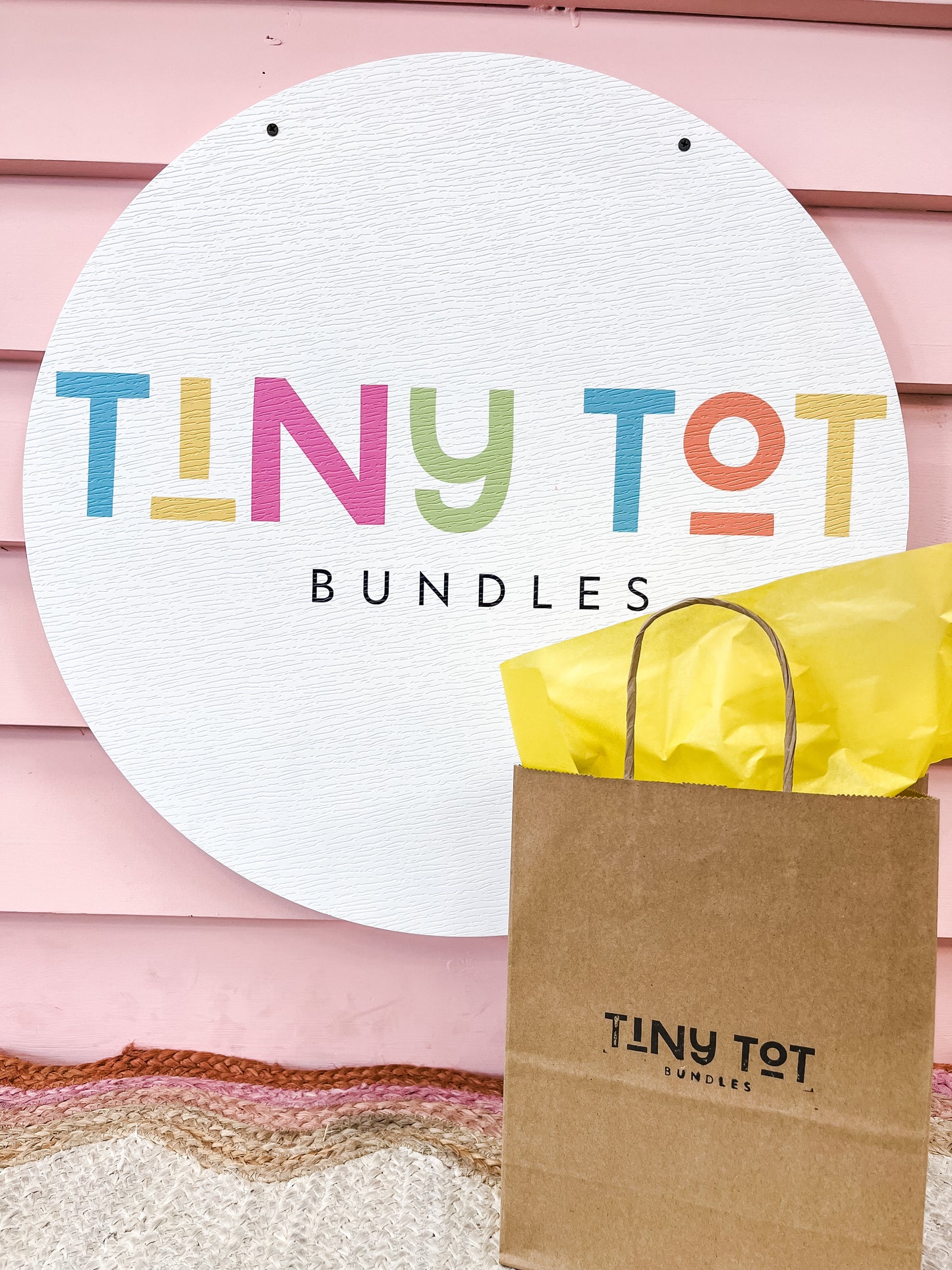 A brown paper bag labeled "Mystery Bundle" from Tiny Tot Bundles sits in front of a round sign with the colorful logo, against a pink wall. Yellow tissue paper peeks out, hinting at surprises and fun activities inside.