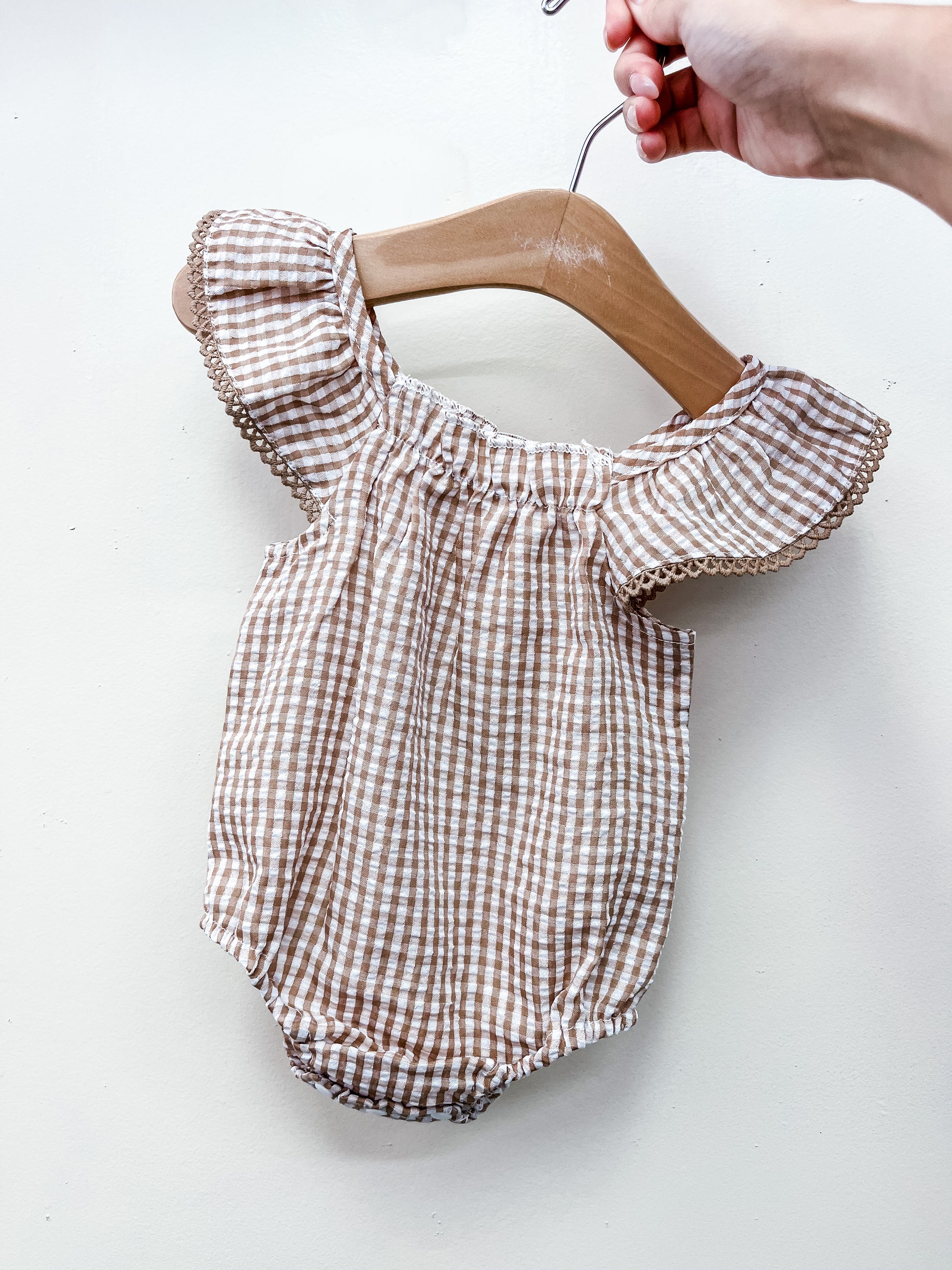 A hand holds the Brown Gingham Dainty Romper by Tiny Tot Bundles—a baby romper with brown and white gingham pattern, ruffled sleeves, and decorative trim—displayed on a wooden hanger against a plain white background.